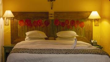 Une chambre d’hôtel avec un lit, deux lampes fixées au mur, une tête de lit en bois ornée de motifs floraux, une croix accrochée au mur et un téléphone sur la table de chevet.