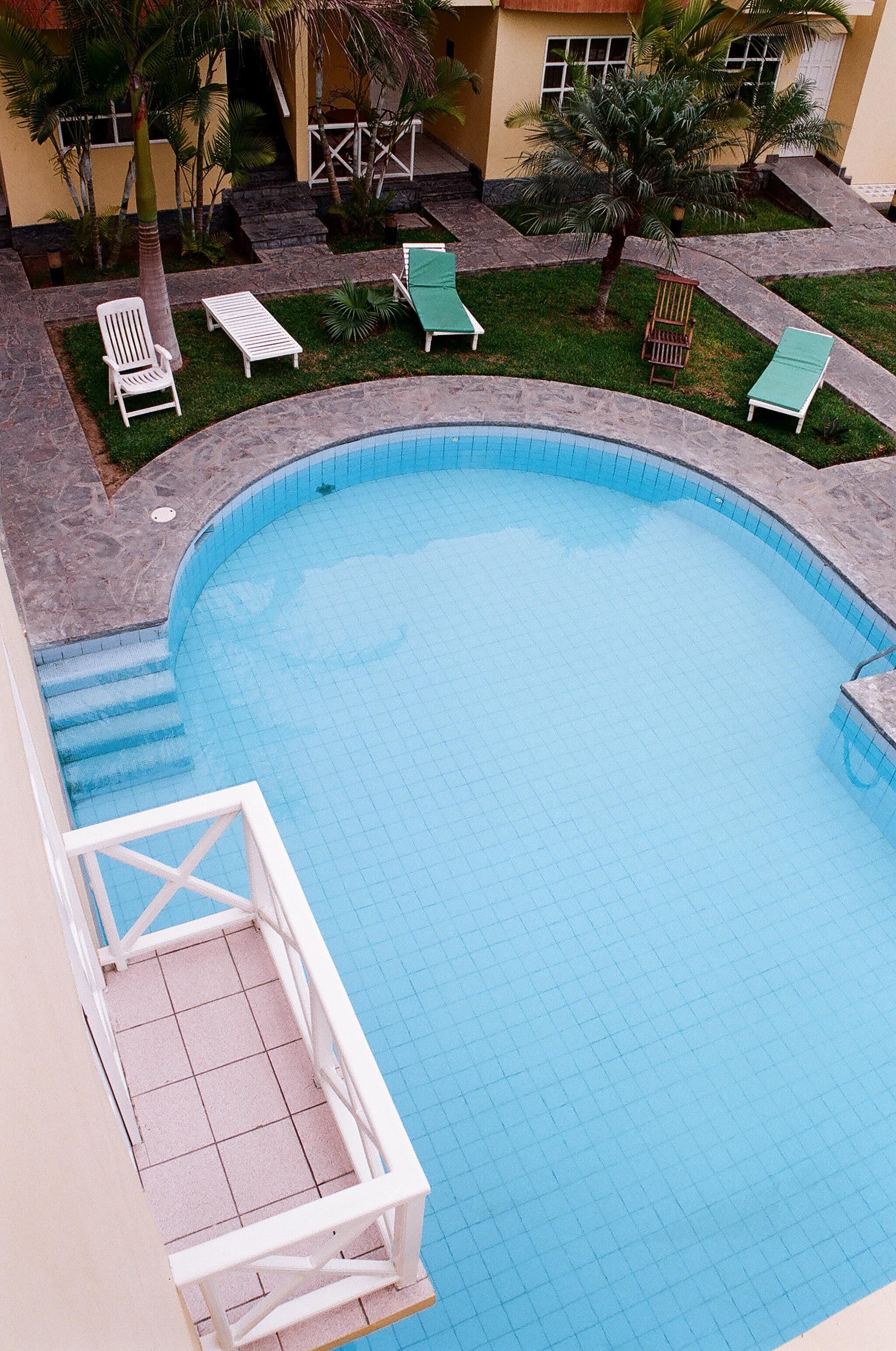 Una piscina con bordes azulejados, rodeada de sillones blancos y un pequeño jardín con palmeras.