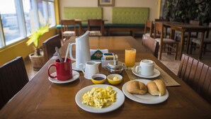 Mesa de café da manhã com pratos de ovos, torradas e suco, em um quarto com assentos estofados em verde e vista através de grandes janelas.