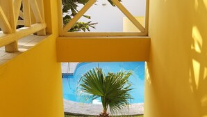 Corredor amarelo que leva a uma área de piscina, com uma palmeira e um edifício azul ao fundo.