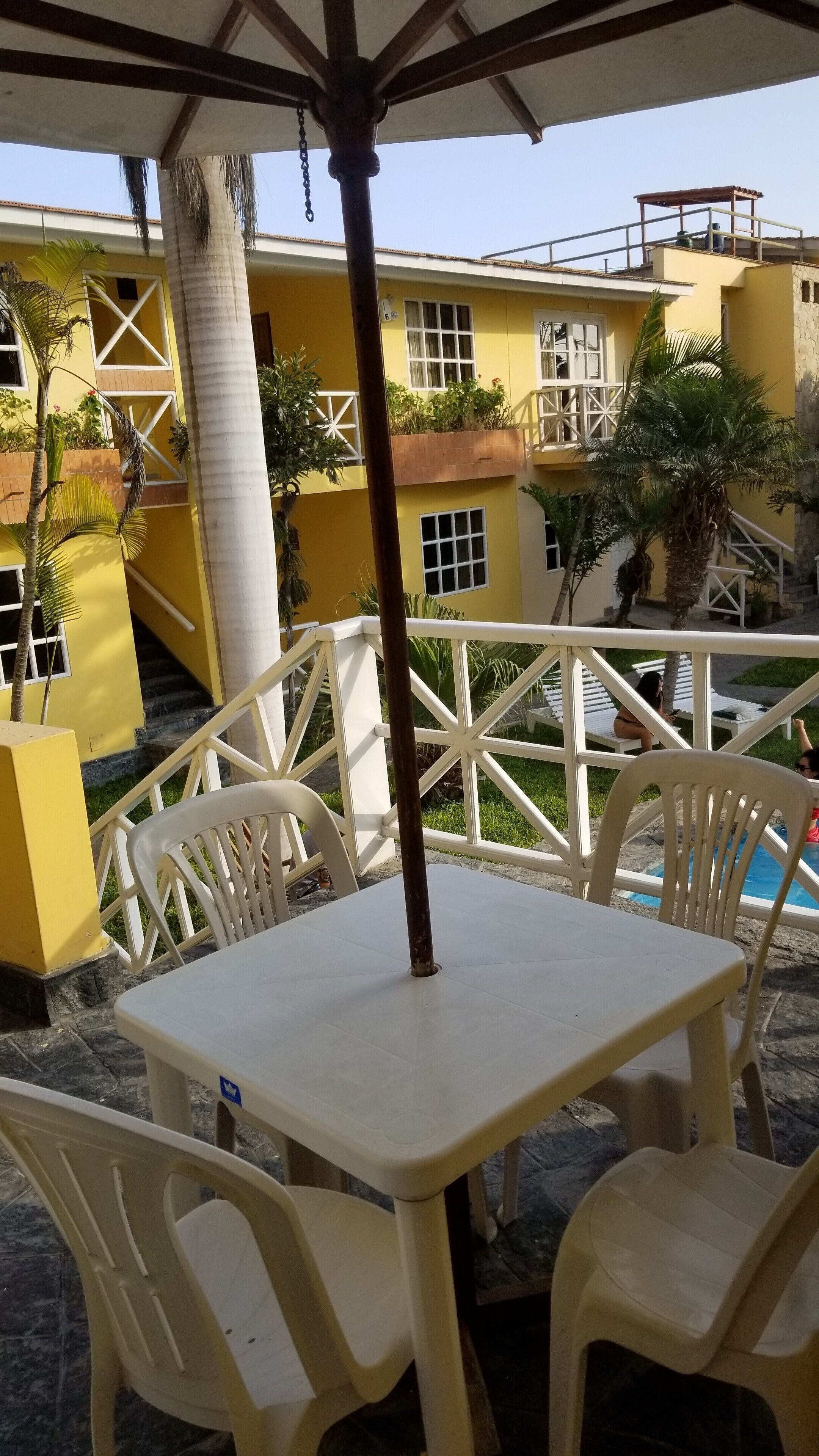An outdoor seating area with a table and chairs, a pool, and a yellow building with balconies.