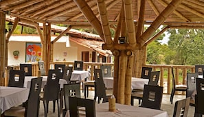Un espacio de comedor al aire libre con estructuras de bambú, mesas con manteles blancos y sillas negras.