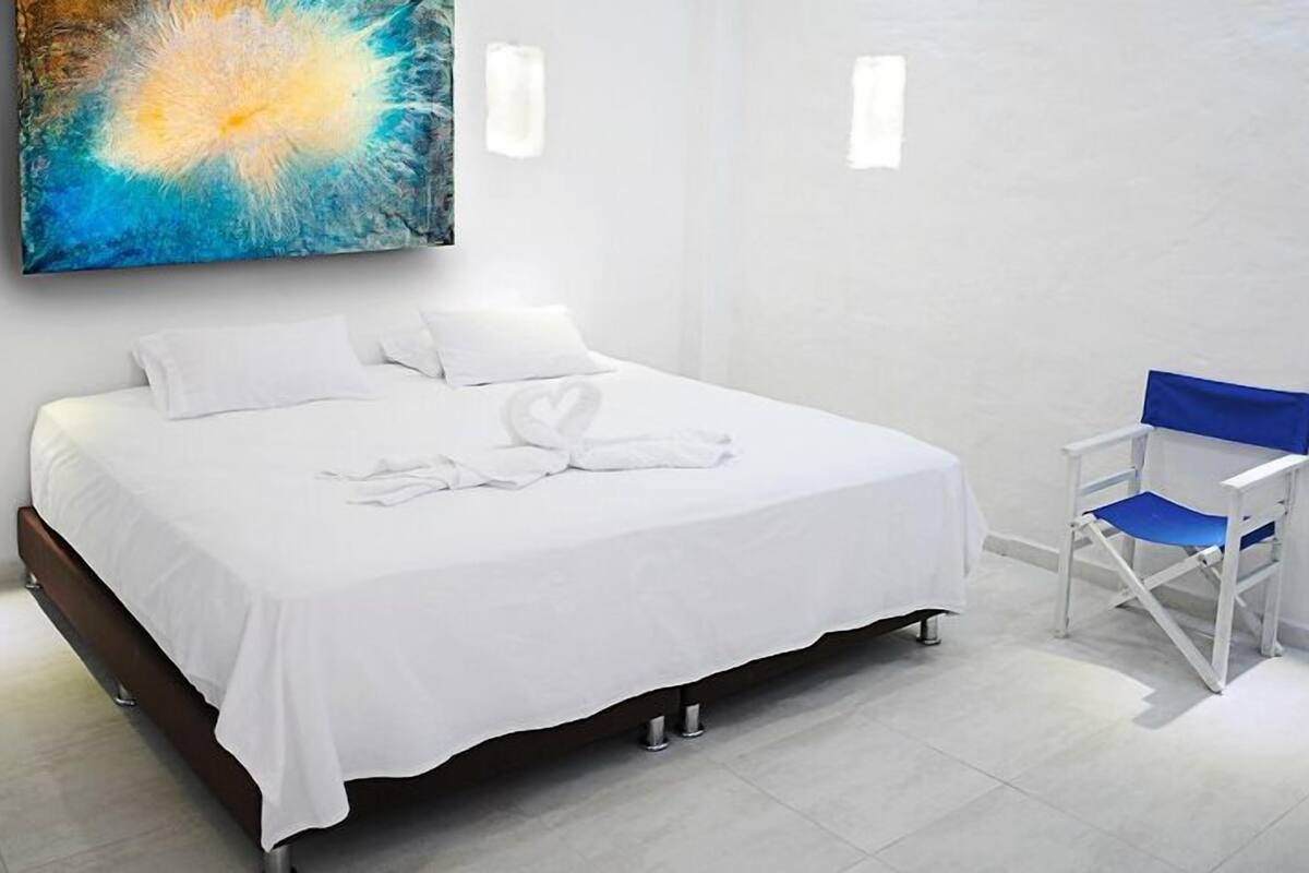 Una habitación de hotel moderna con una cama bien hecha, una silla plegable azul y un cuadro abstracto grande en la pared.