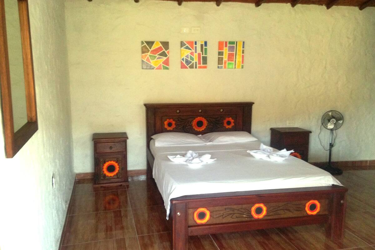 Un dormitorio con una cama de madera, dos mesitas de noche, un ventilador de pared y tres obras de arte coloridas.