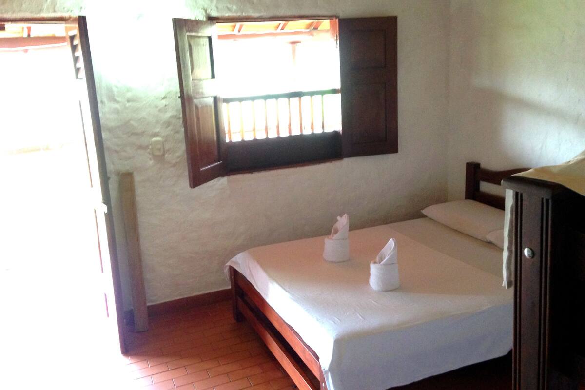 Una habitación con una cama de madera, un ventanal con barrotes y una pared blanca.