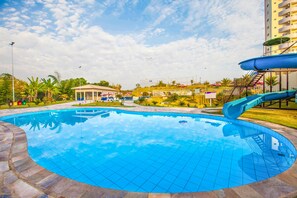 A swimming pool with a slide, surrounded by a grassy area and palm trees. - Golden Dolphin Express OFICIAL (Caldas Novas)