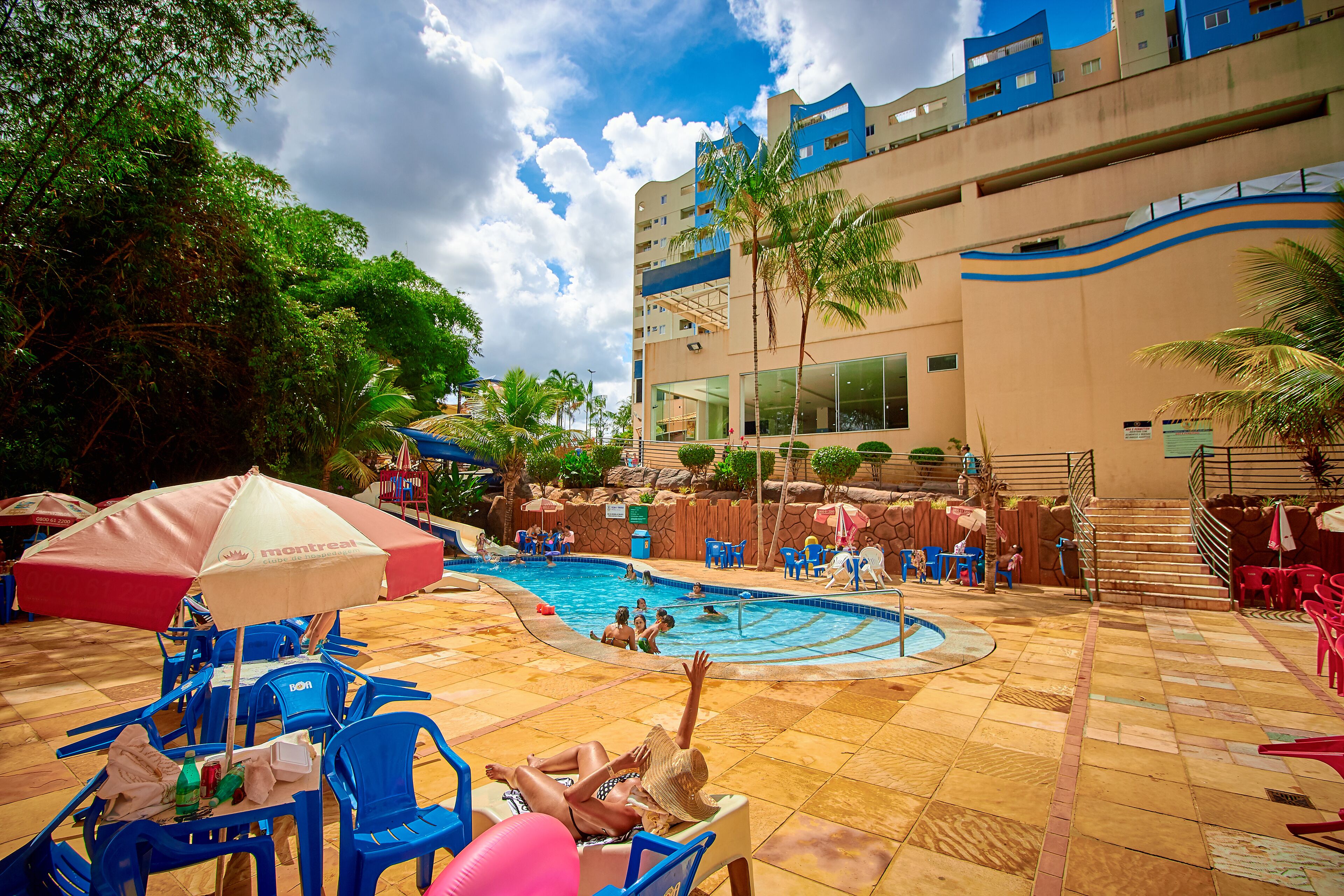 A pool area with blue chairs and umbrellas, a swimming pool, and a building with balconies.