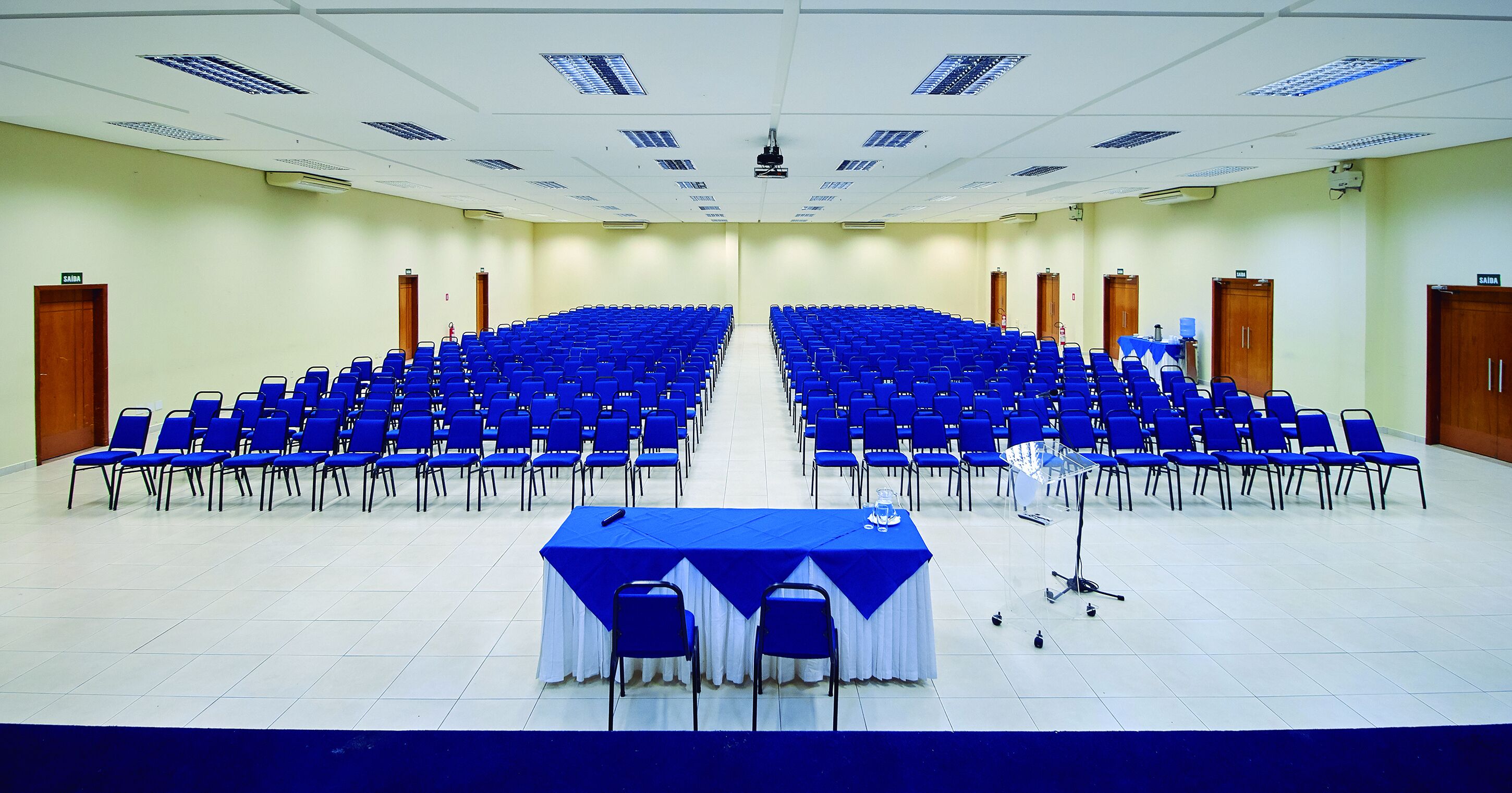 A large conference room with rows of blue chairs, a central table with a white tablecloth, and a projector screen at the front.