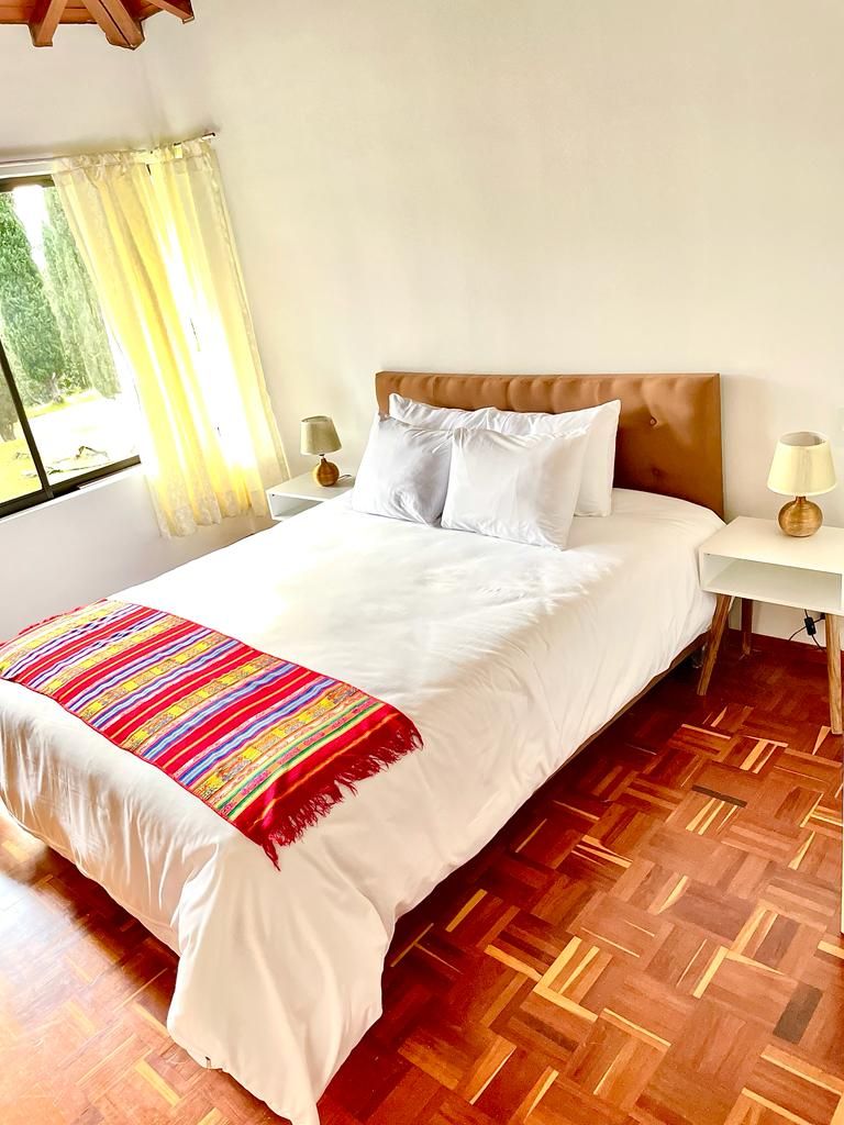 Un lit bien fait, avec du linge de lit blanc et une couverture rayée aux couleurs vives, deux tables de chevet avec des lampes, et un plancher en parquet.