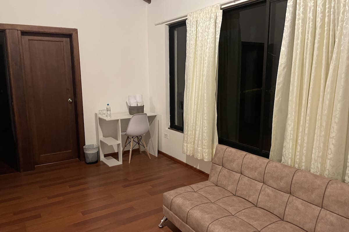 Une pièce avec un plancher en bois, un canapé, un bureau avec une chaise, une fenêtre avec des rideaux et une porte.