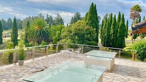 A rooftop terrace with glass tables and chairs, surrounded by a garden with various trees and a view of the countryside.