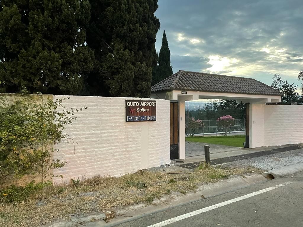 Ein Gebäude mit der Aufschrift „Quito Airport Suites“, einem überdachten Eingang und einem Parkschild, das eine rund um die Uhr Zufahrt anzeigt.