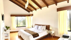 A spacious bedroom with a large bed, wooden ceiling beams, a bedside table with a lamp, and a view of the outdoors through a window.