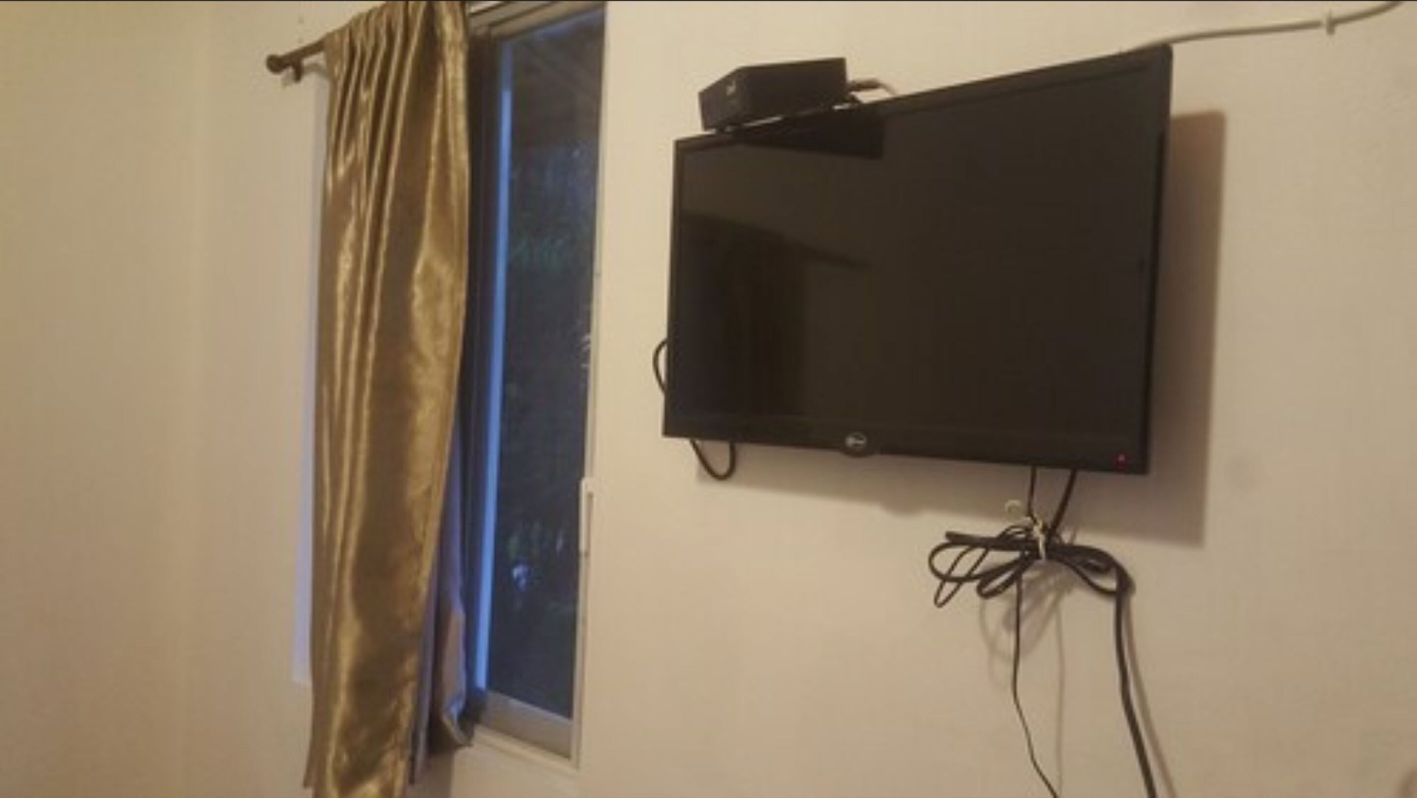 A room with a mounted flat-screen TV on the wall, a window with a curtain, and visible electrical cords.