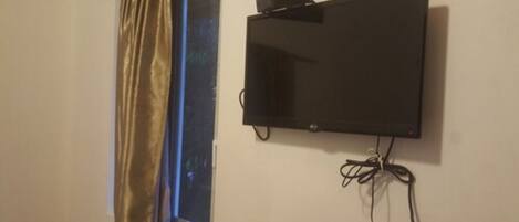 A room with a mounted flat-screen TV on the wall, a window with a curtain, and visible electrical cords.