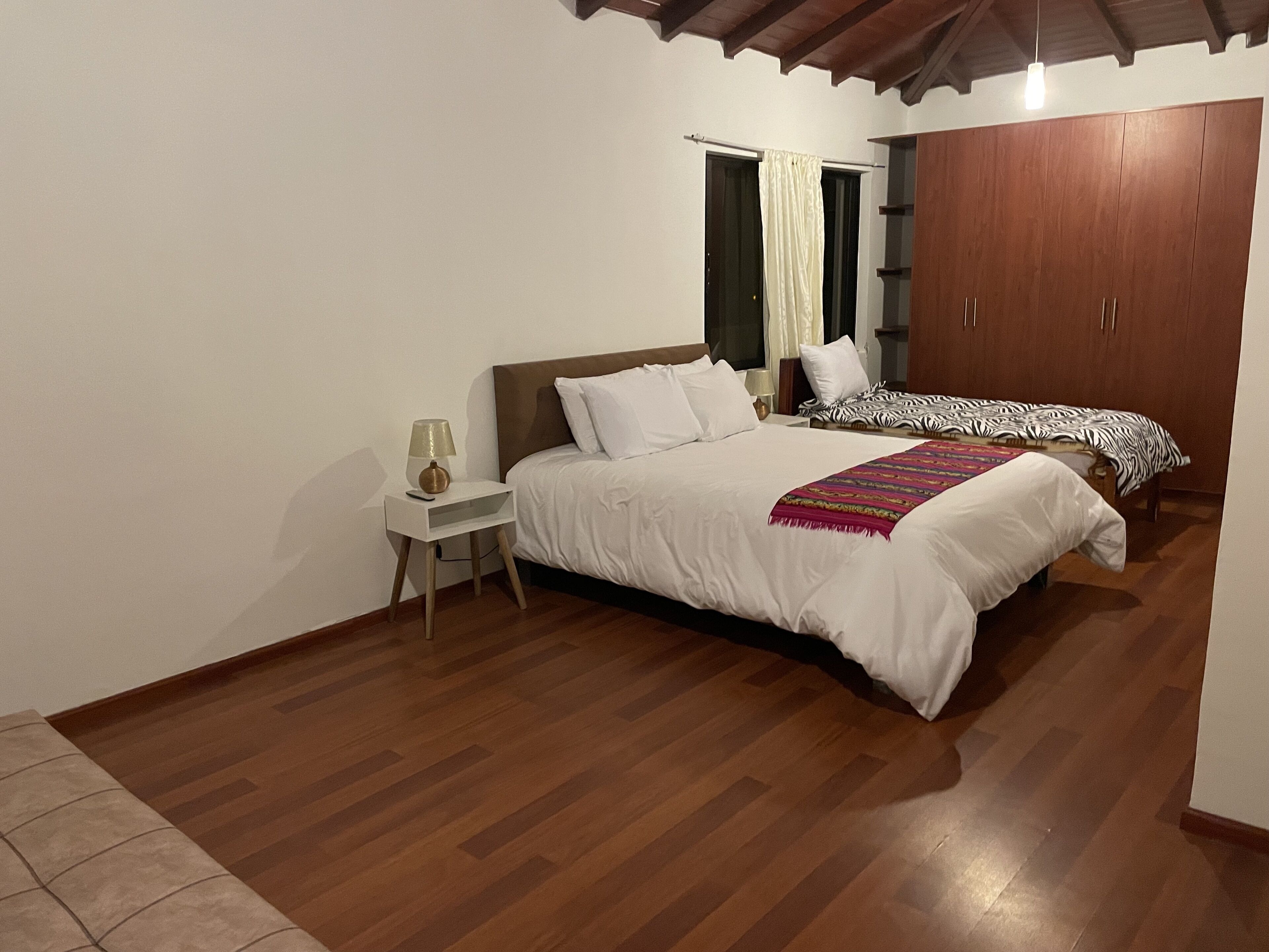 A bedroom with a large bed, a smaller bed, a nightstand with a lamp, wooden flooring, and a wardrobe.