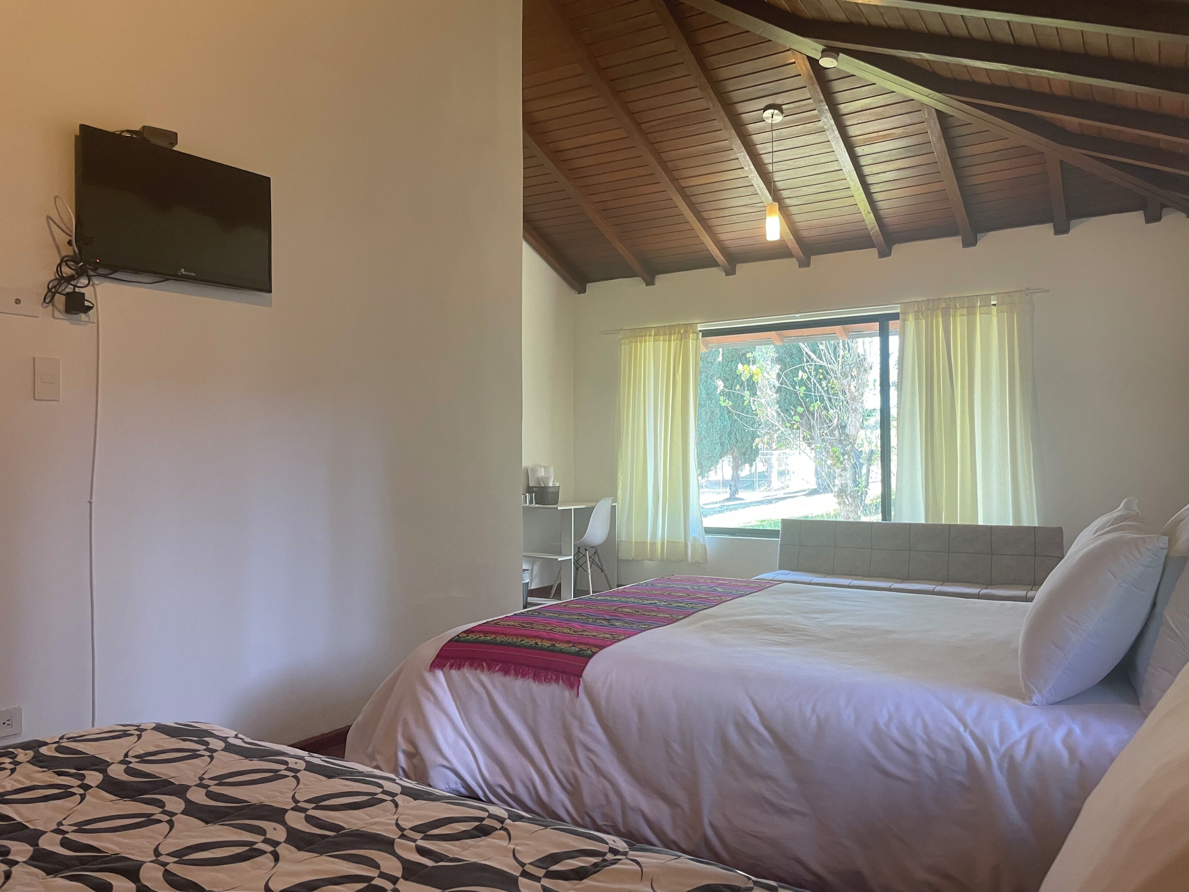Un dormitorio con dos camas, un televisor montado en la pared, una ventana con cortinas y un techo con vigas de madera.