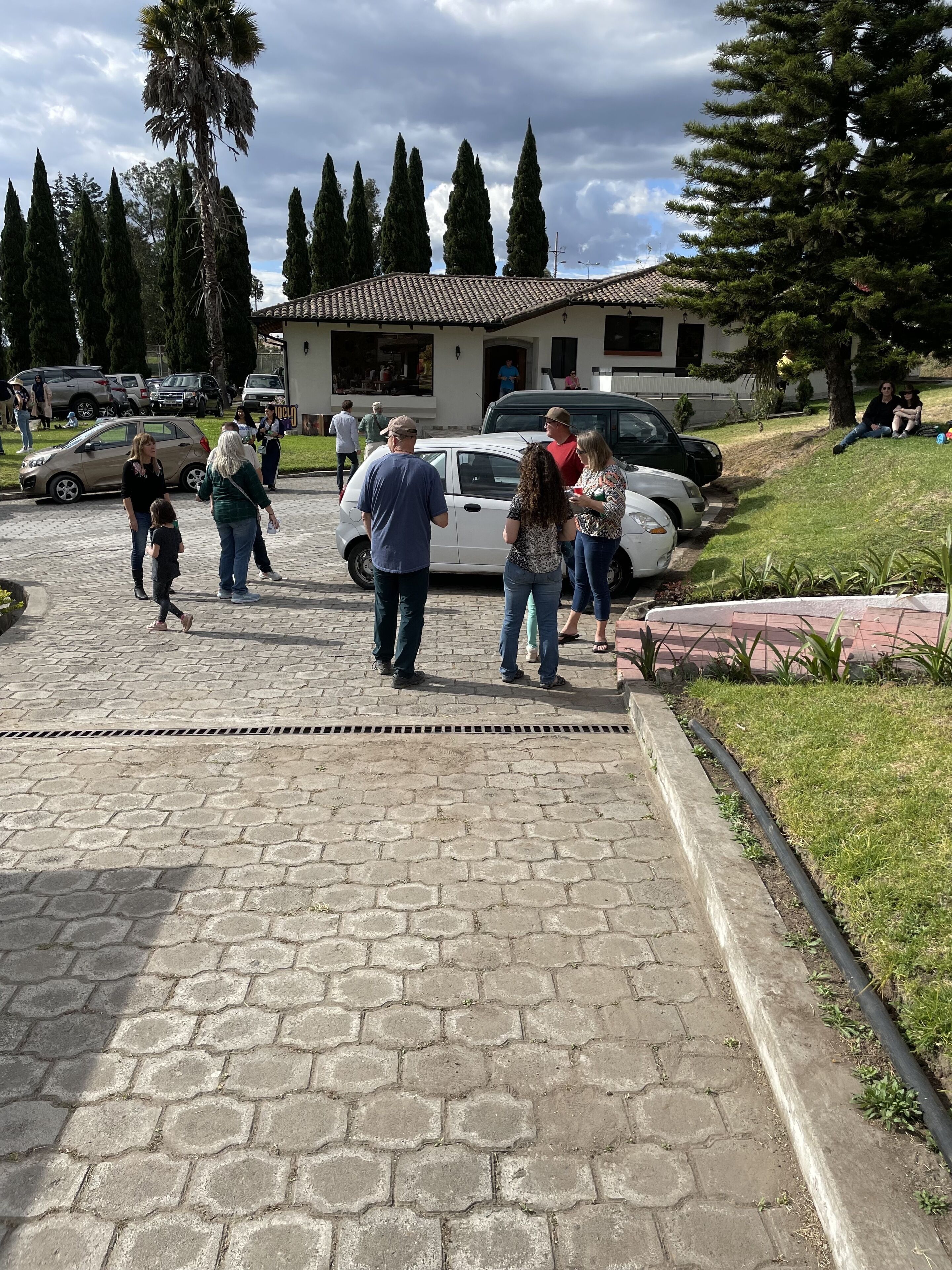 A paved pathway leading to a building with a tiled roof, surrounded by parked cars and people.