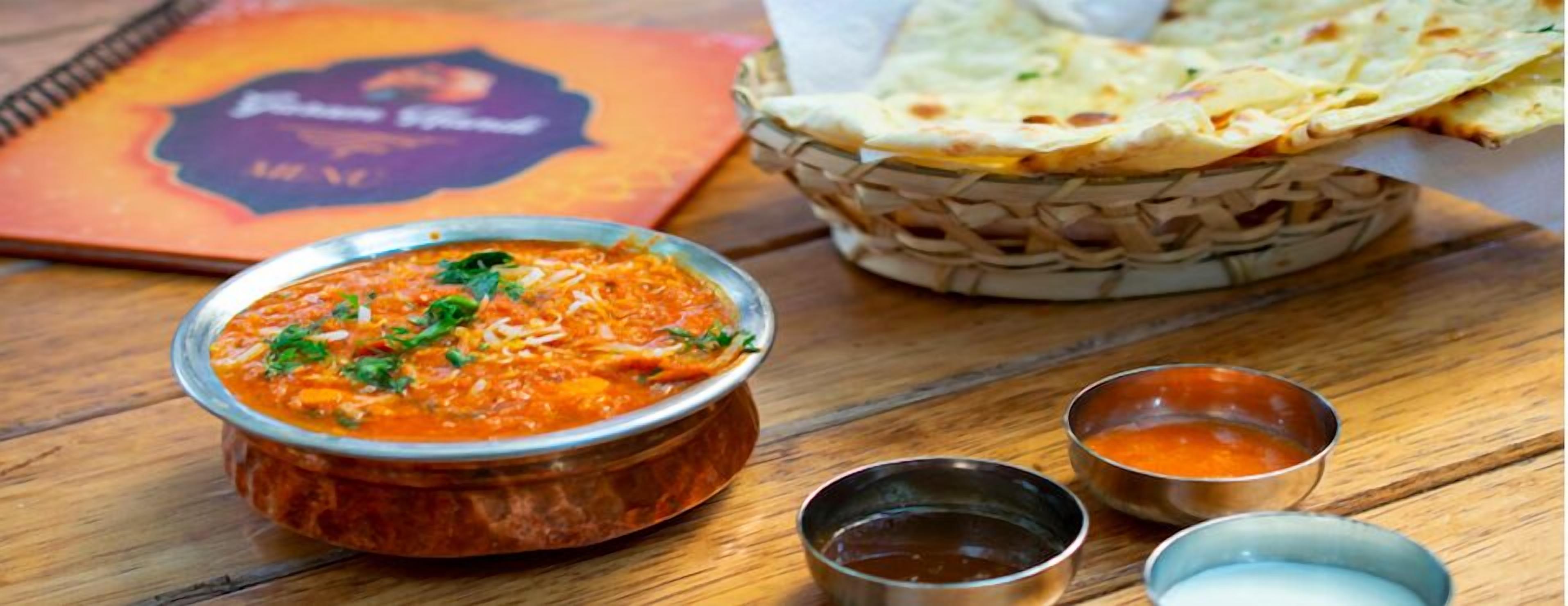 A meal with a bowl of curry, naan bread, and two small bowls of sauces on a wooden table.