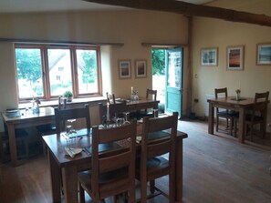 A dining area with wooden tables and chairs, a window with a view of greenery, and framed pictures on the wall.