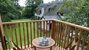 Terraza de madera con una mesa dispuesta para el té, rodeada de sillas, con vistas a una casa de techo de paja y abundante vegetación.