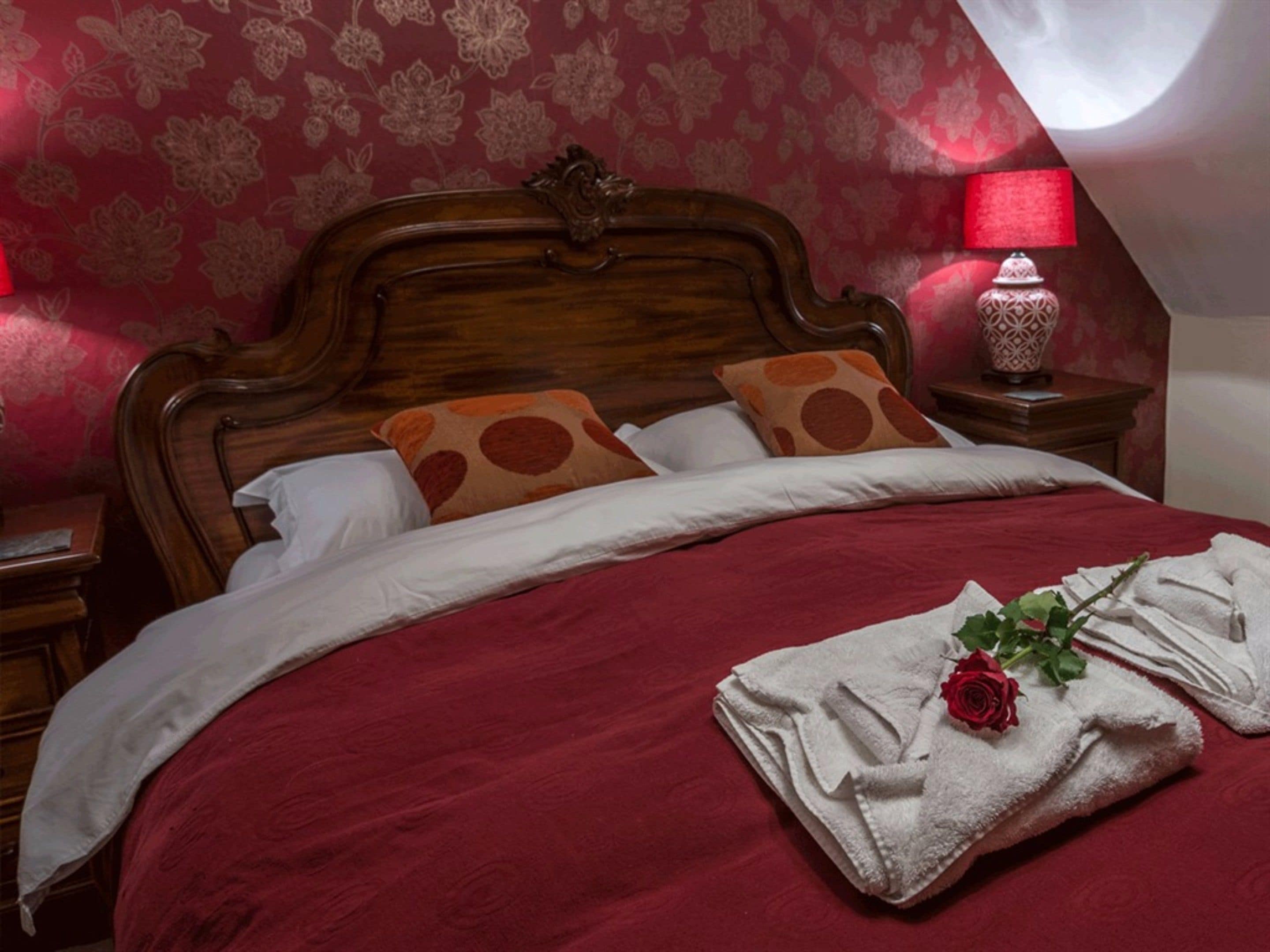 Una cama bien tendida con una colcha roja, almohadas blancas y una almohada decorativa con lunares naranjas. Hay una mesita de noche con una lámpara y un jarrón decorativo. Una rosa está colocada sobre una toalla doblada en la cama.