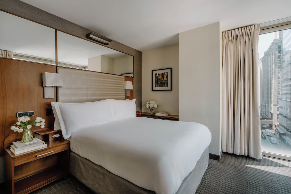 Una habitación de hotel con una cama grande, una mesita de noche con una lámpara, un jarrón con flores y una ventana con cortinas.