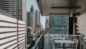 Terraço no telhado com mesas e cadeiras, oferecendo vista dos edifícios altos ao redor.