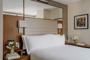 A neatly made hotel room with a large bed, a bedside table with a lamp, a mirror, and a framed picture on the wall.