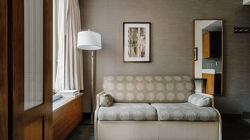 A modern hotel room with a sofa, a standing lamp, a framed picture, and a mirror reflecting a bathroom.