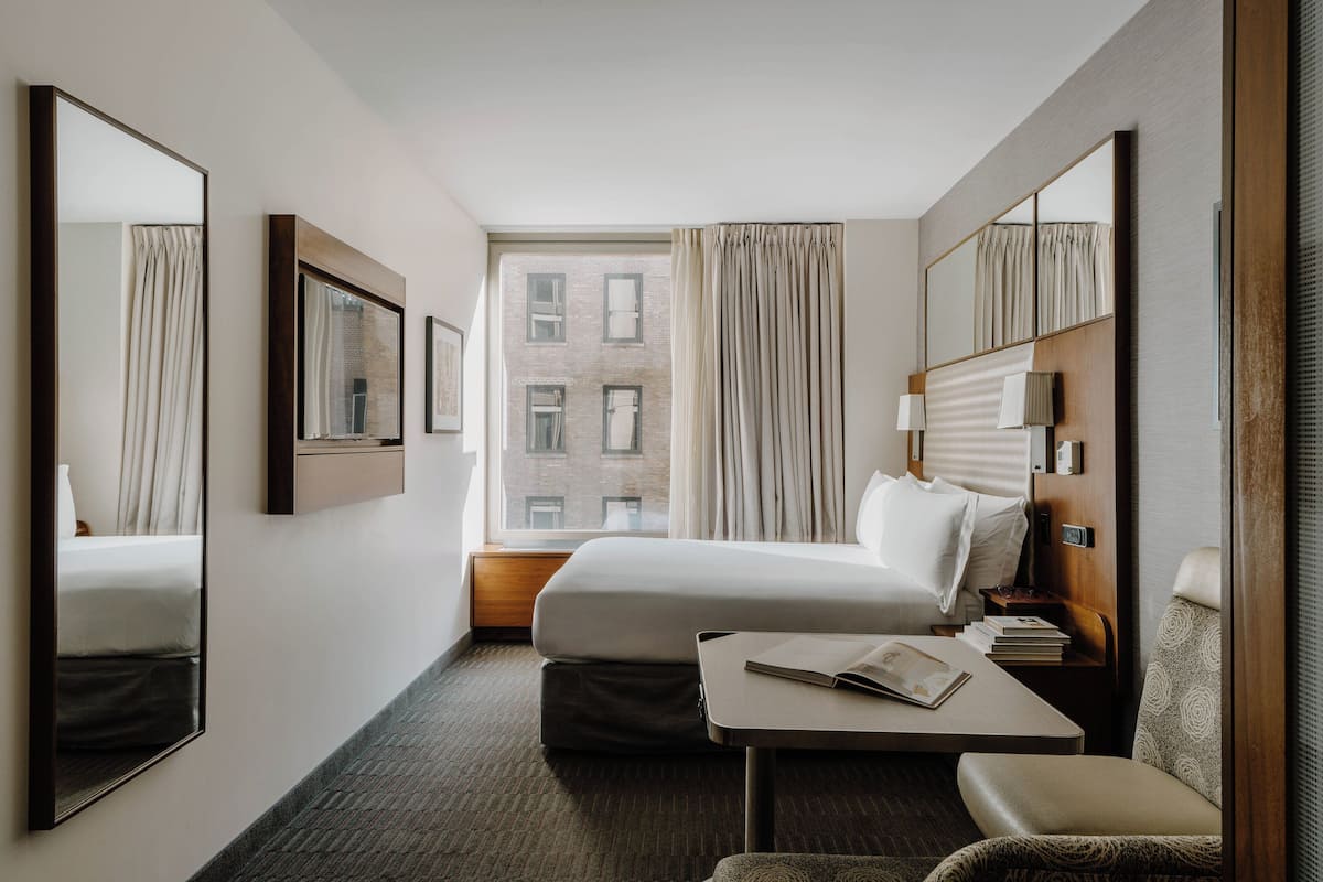 Habitación de hotel con una cama grande, una mesita de noche con lámpara, una silla y un ventanal con cortinas.