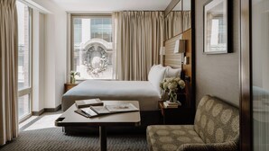 Quarto de hotel com uma cama grande, uma mesa de cabeceira com abajur, uma cadeira e vista para a paisagem urbana pela janela.
