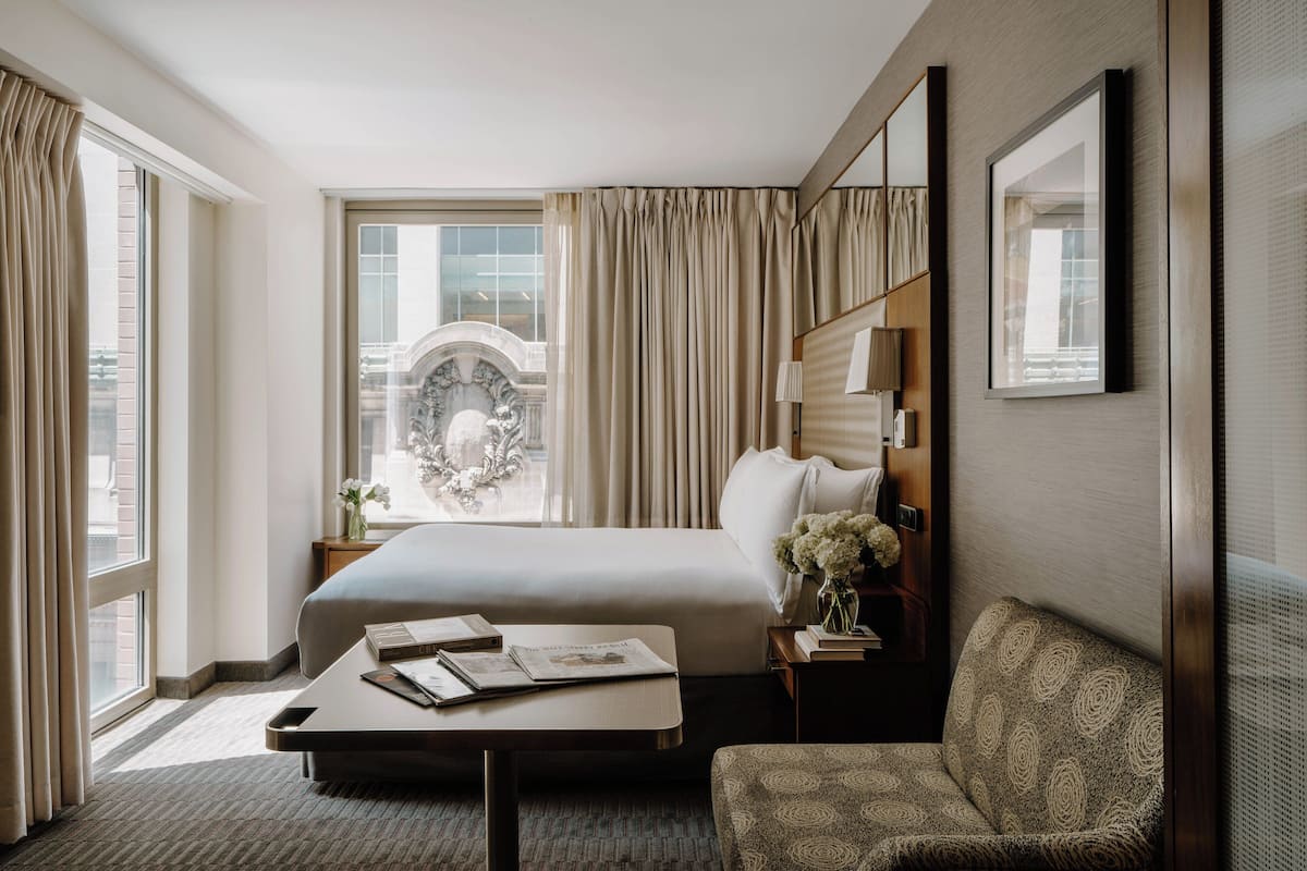 Habitación de hotel con una cama grande, una mesita de noche con lámpara, una silla y vista a un paisaje urbano a través de la ventana.