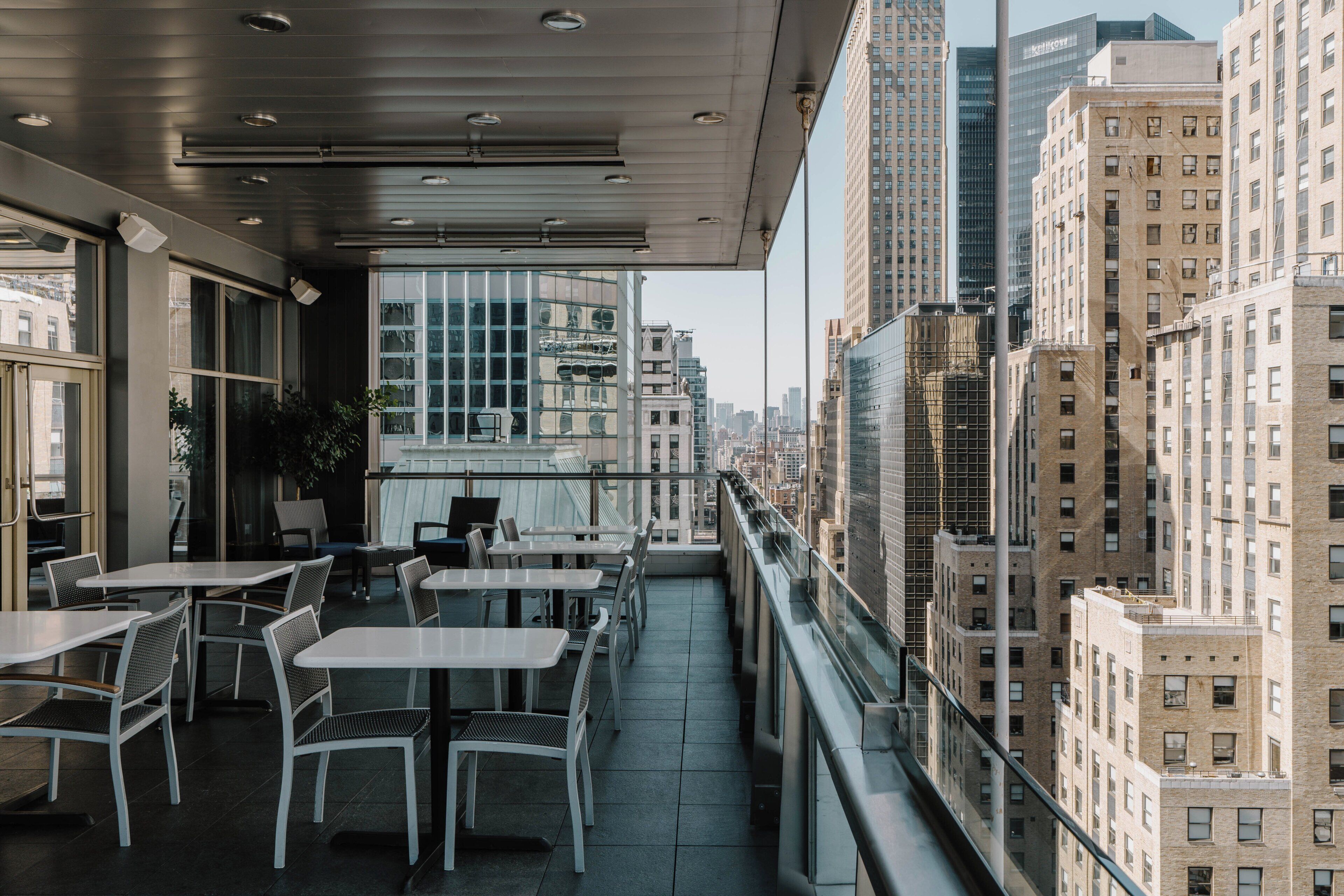 Un área de comedor al aire libre con mesas y sillas, con vista a un paisaje urbano con edificios altos.