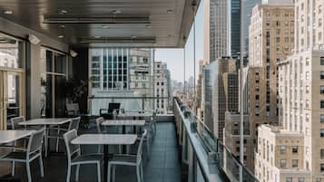 Un área de comedor al aire libre con mesas y sillas, con vista a un paisaje urbano con edificios altos.