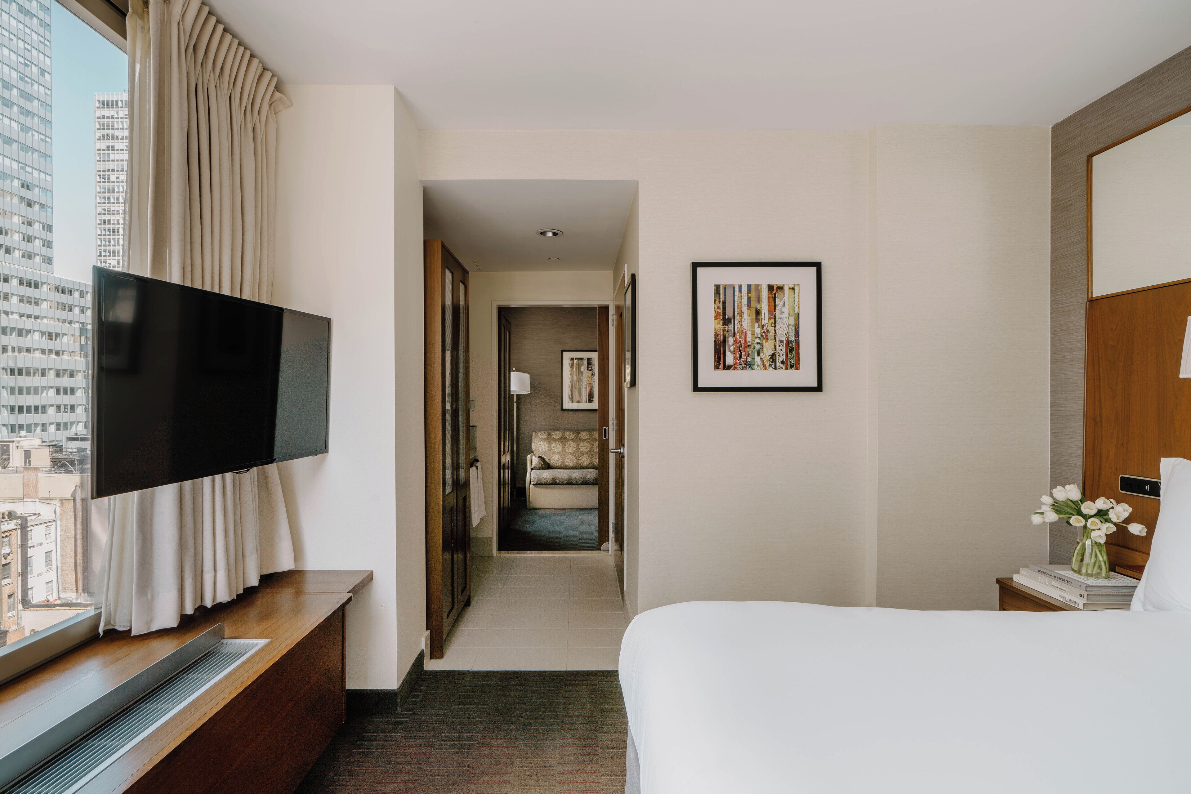 A hotel room with a bed, a TV on a stand, a desk with a computer, a window with curtains, and a hallway leading to another room.