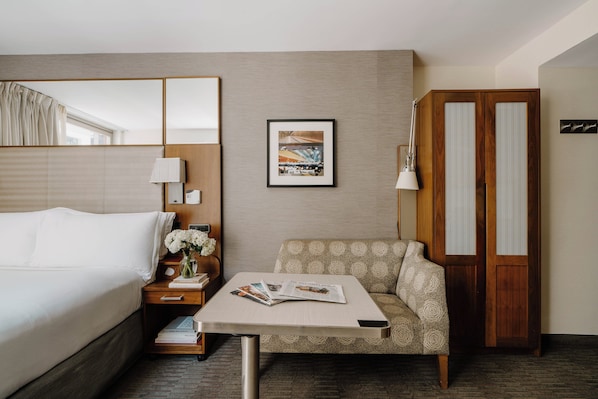 Quarto de hotel com cama, mesa de cabeceira com luminária, pequena mesa com revista, poltrona estampada e guarda-roupa.