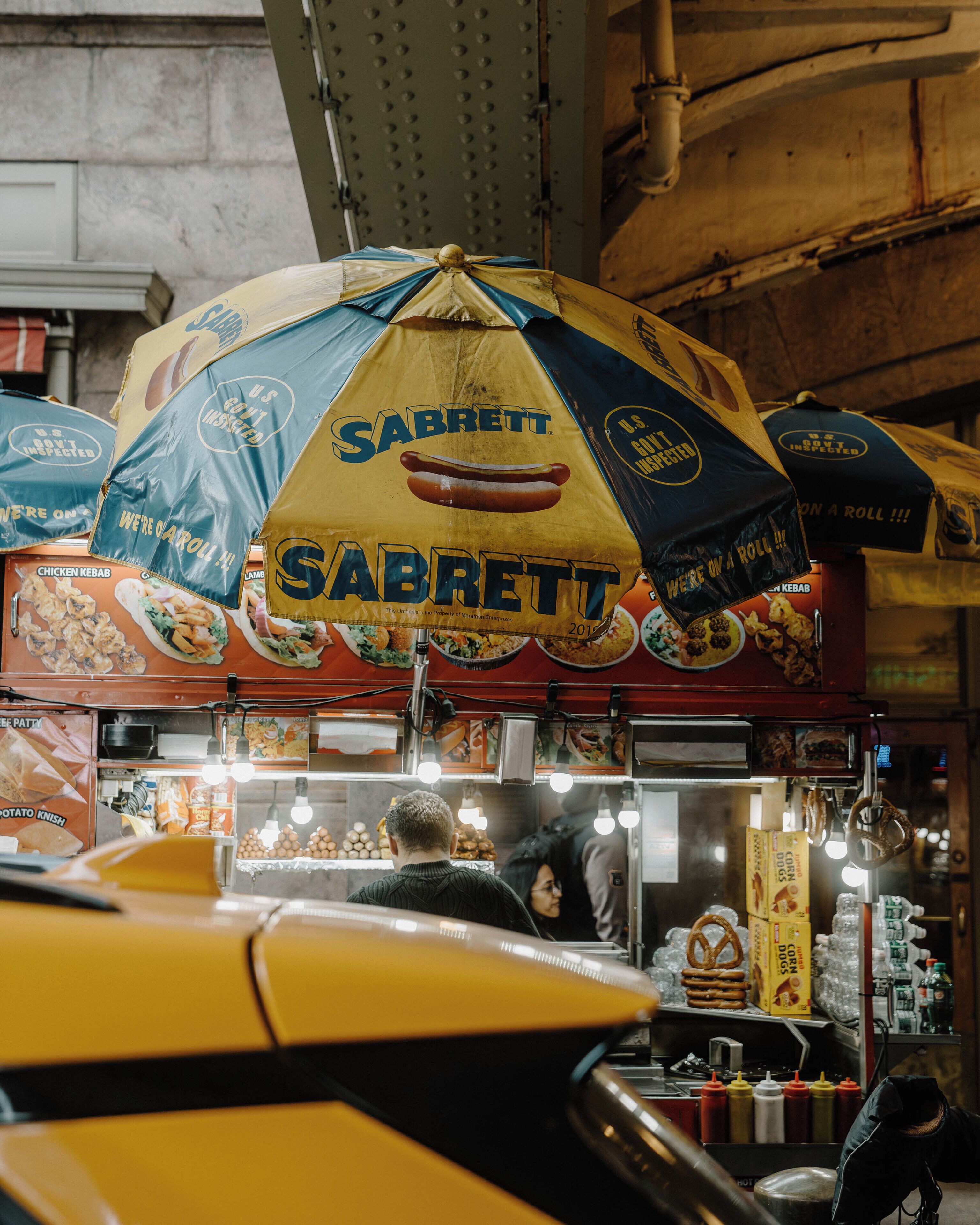 Un carrito de comida con un paraguas grande que dice 'Sabrett' y diferentes productos alimenticios exhibidos debajo.