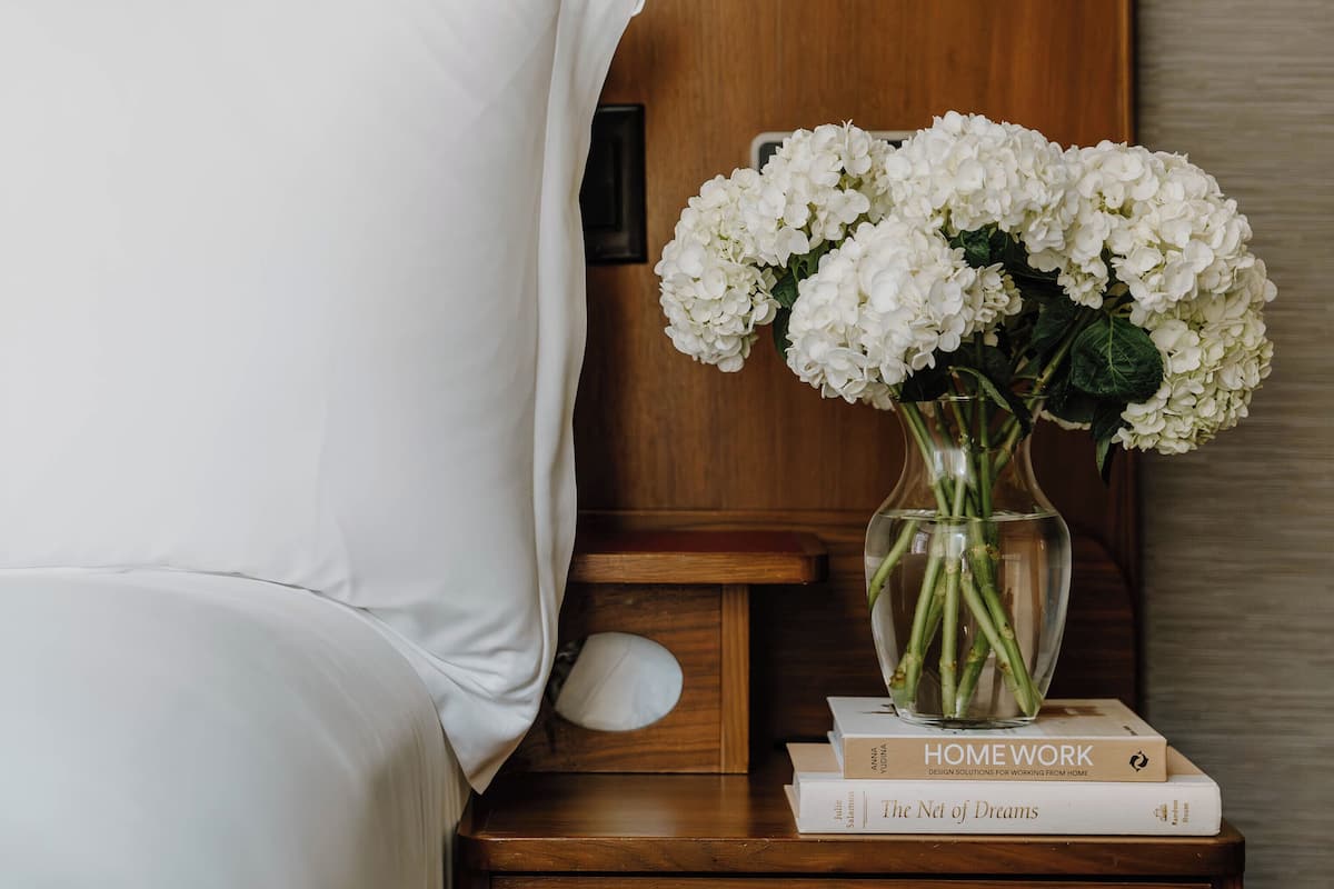 Una cama bien hecha con sábanas blancas, una mesita de noche con un florero de flores blancas, y dos libros titulados 'HOMEWORK' y 'The Net of Dreams'.