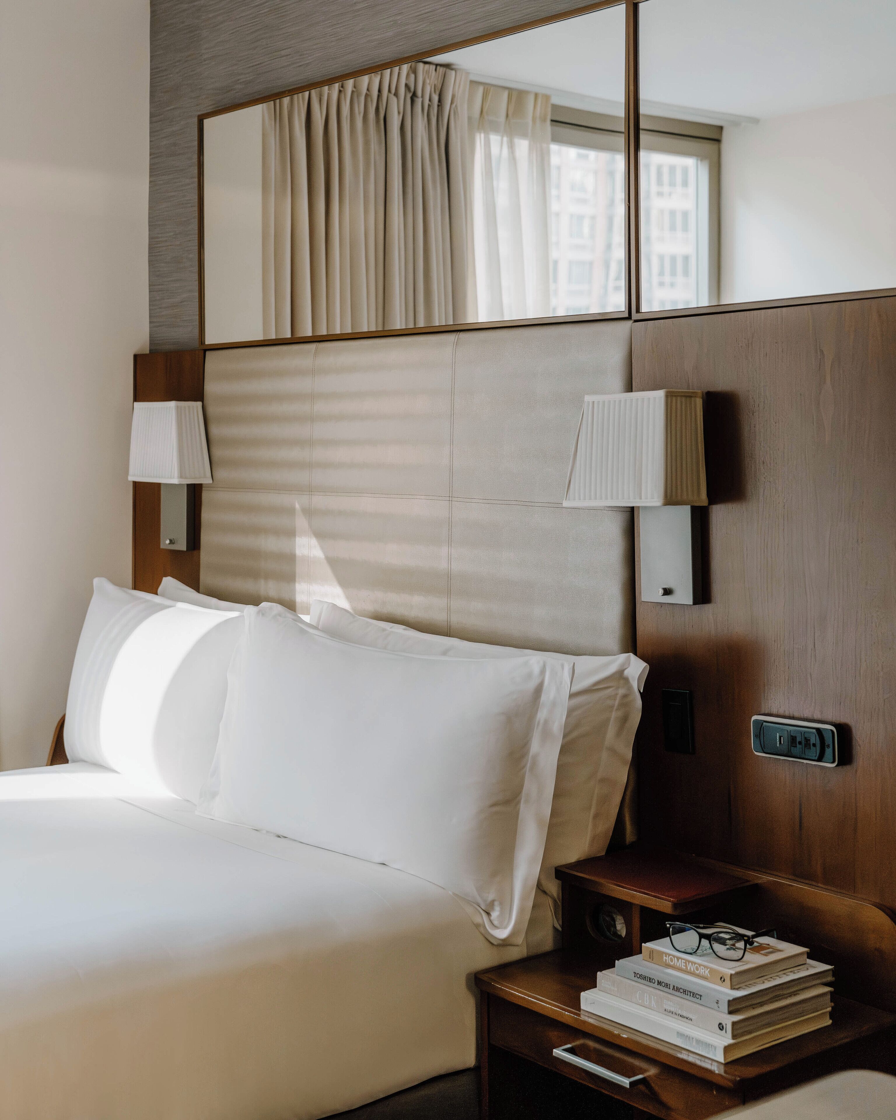 A hotel room with a large bed, two bedside lamps, a nightstand with books and glasses, and a mirror above the bed.