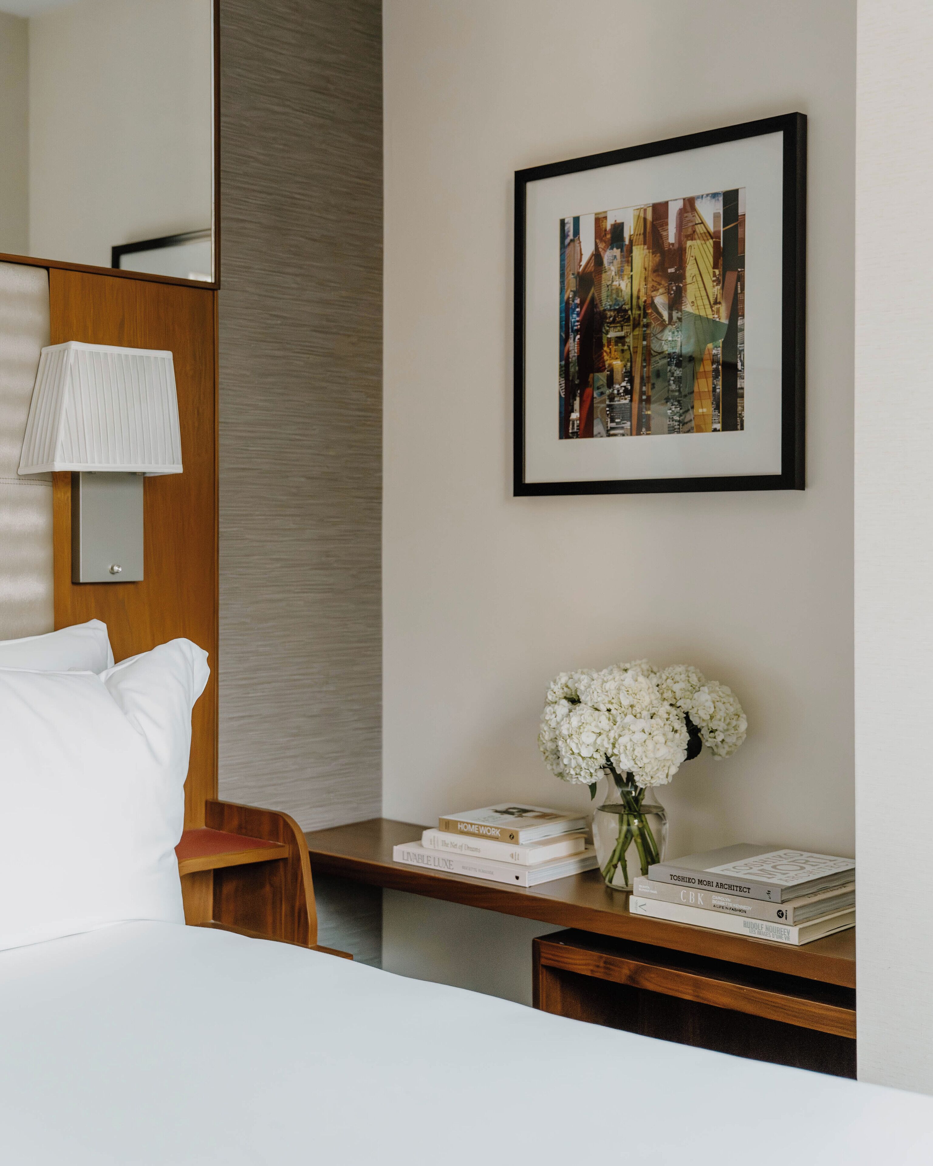 Habitación de hotel con cama, mesita de noche con libros y un jarrón de flores, lámpara y una obra de arte con una vista urbana enmarcada en la pared.