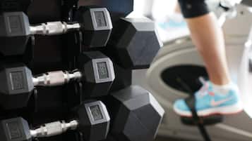 A close-up of a set of black hexagonal dumbbells with weight markings, and a person's foot in a blue athletic shoe on a treadmill.