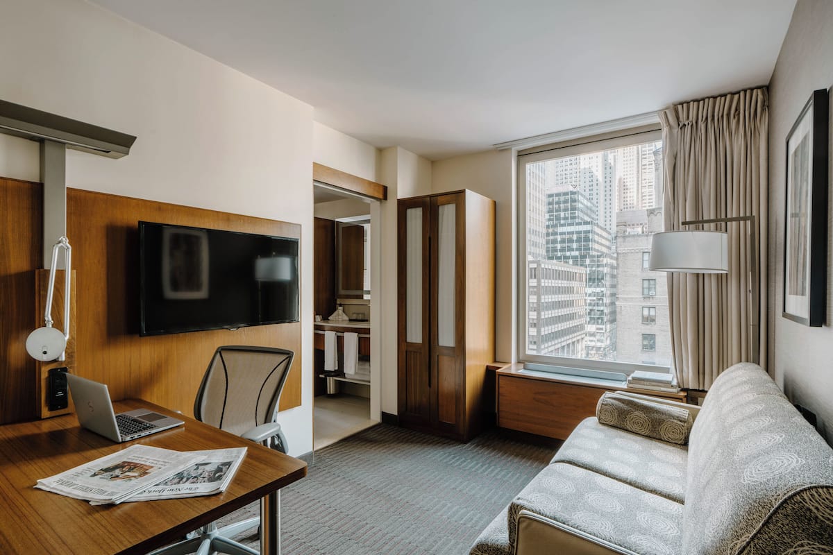 Habitación de hotel con escritorio de madera, una laptop, un televisor de pantalla plana, un sofá y un ventanal con vista a la ciudad.