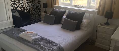 A neatly made bed with a grey duvet, multiple pillows, and a small tray with a bottle and glasses. There is a window with curtains, a bedside table with a lamp, and a patterned wallpaper on the wall.