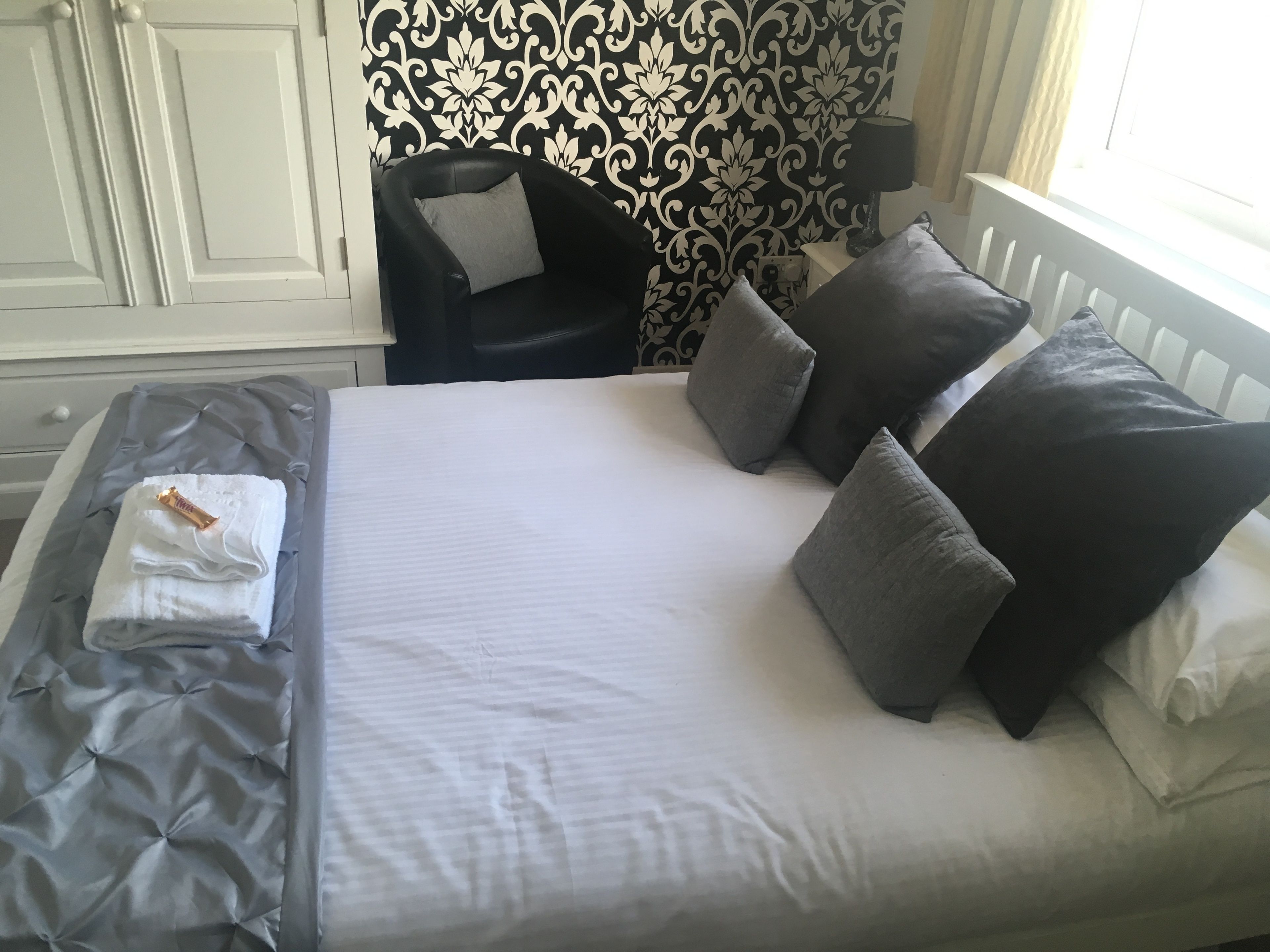 A neatly made bed with grey and white bedding, a black leather chair with a grey pillow, and a patterned wallpaper.