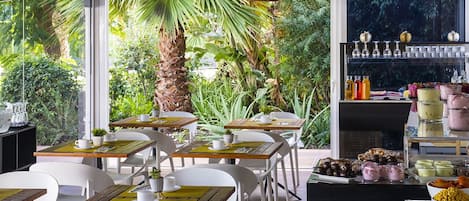 Un comedor interior con mesas y sillas, un bufé con diversas opciones de comida, y vistas a zonas verdes exteriores.