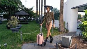 Una persona con un traje de cuerpo entero de leopardo y tacones altos caminando por un sendero pavimentado con una maleta rosa.