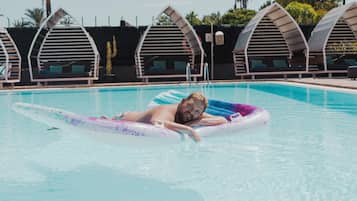 Un hombre recostado en un flotador en una piscina, con sillones de descanso y palmeras al fondo.
