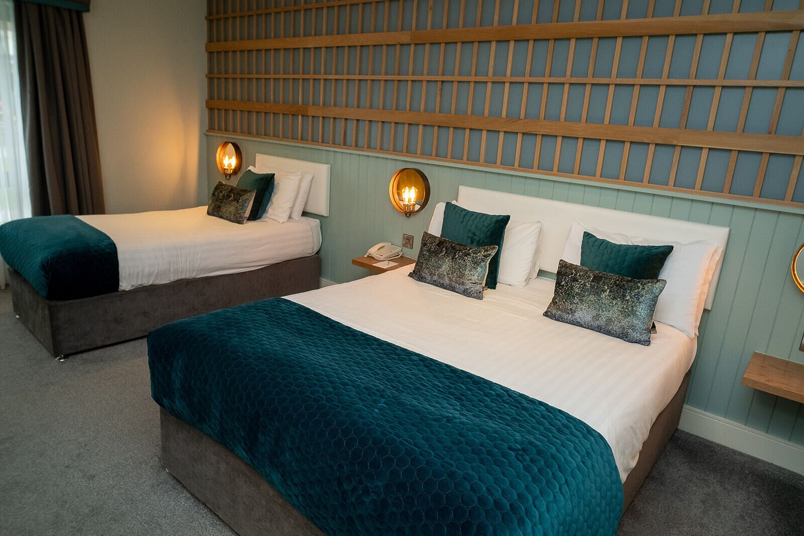A hotel room with two beds, each with a teal bedspread and matching pillows. There are wall-mounted sconces providing light, and a wooden headboard with a grid pattern.