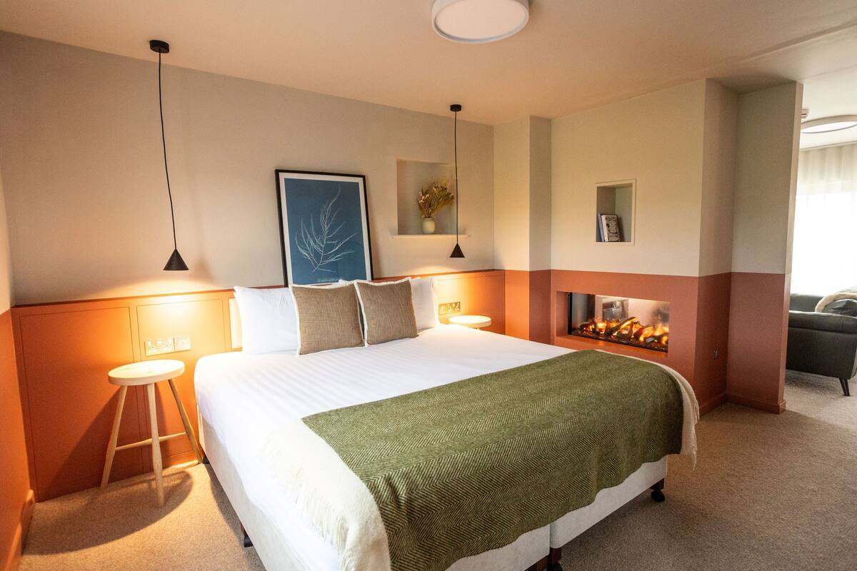 A hotel room with a bed, bedside lamps, a small stool, a wall-mounted shelf with a plant, and a fireplace.